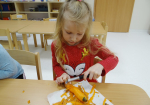 Skupiona Nadia siedzi przy stoliku i obiera marchewkę.